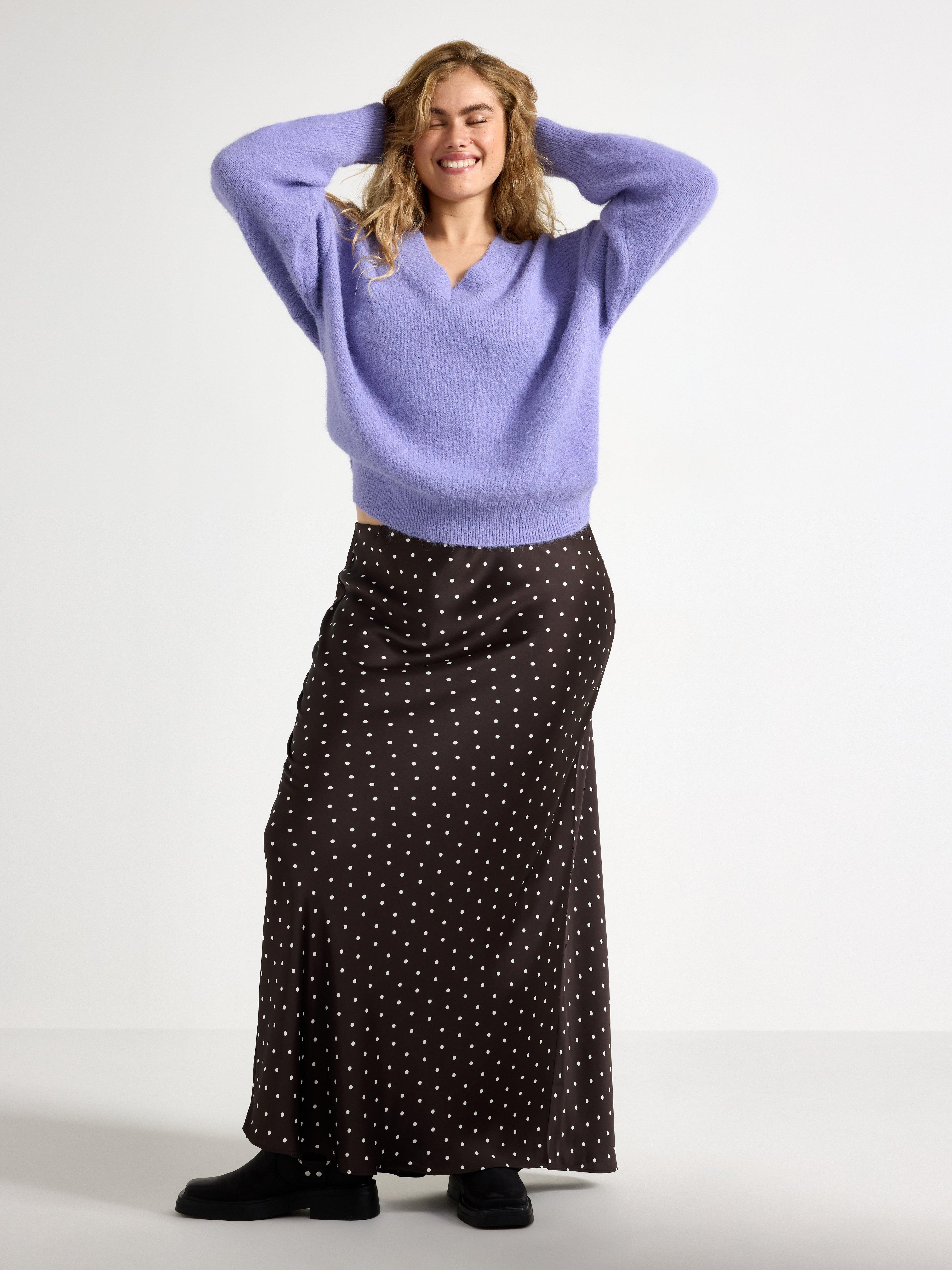  A smiling woman wearing a purple knitted sweater and black and white polka dot skirt.