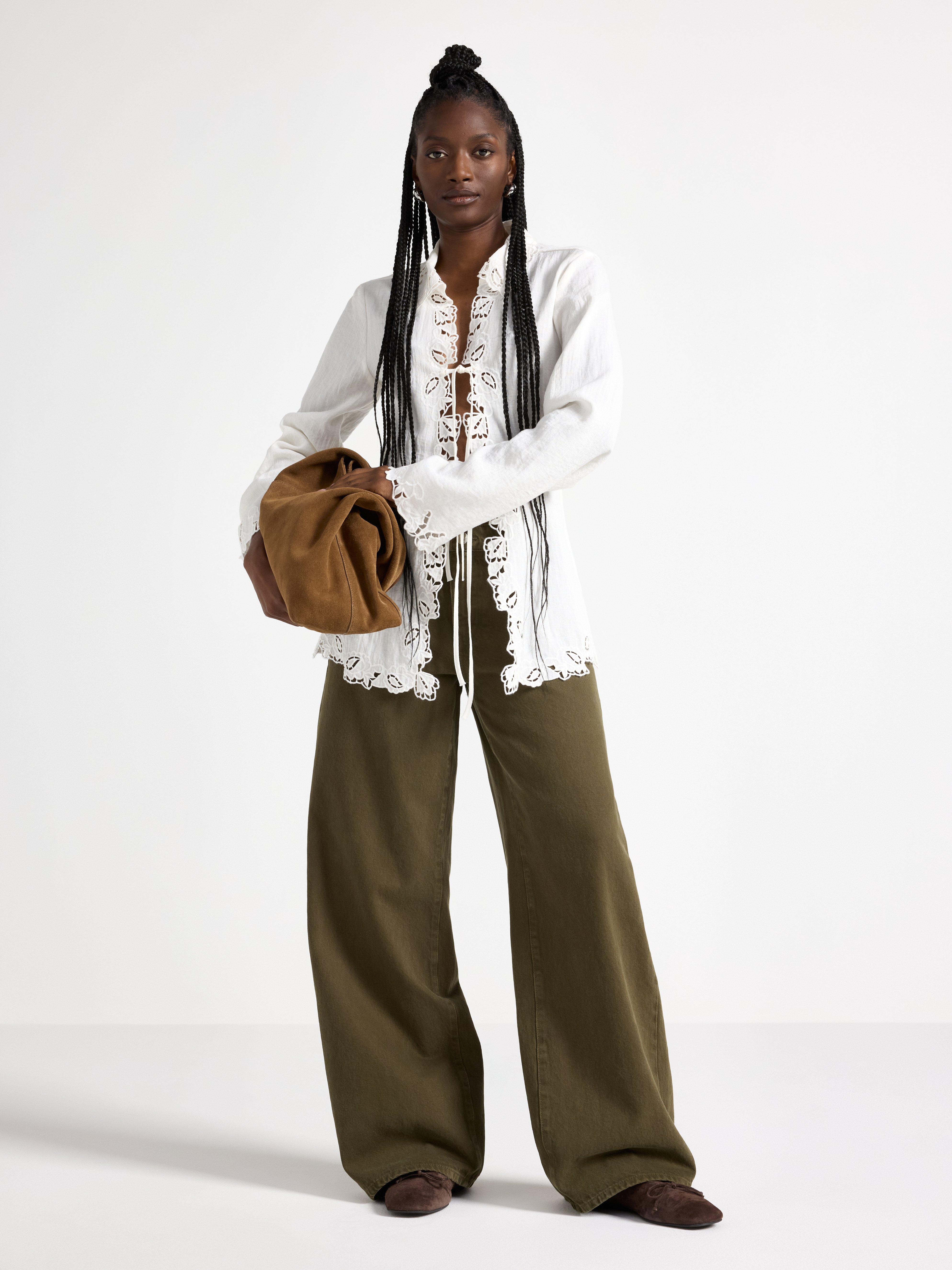  A woman wearing a white embroidered blouse with a tie and a pair of green pants. She is holding a brown shopper bag in her arms.