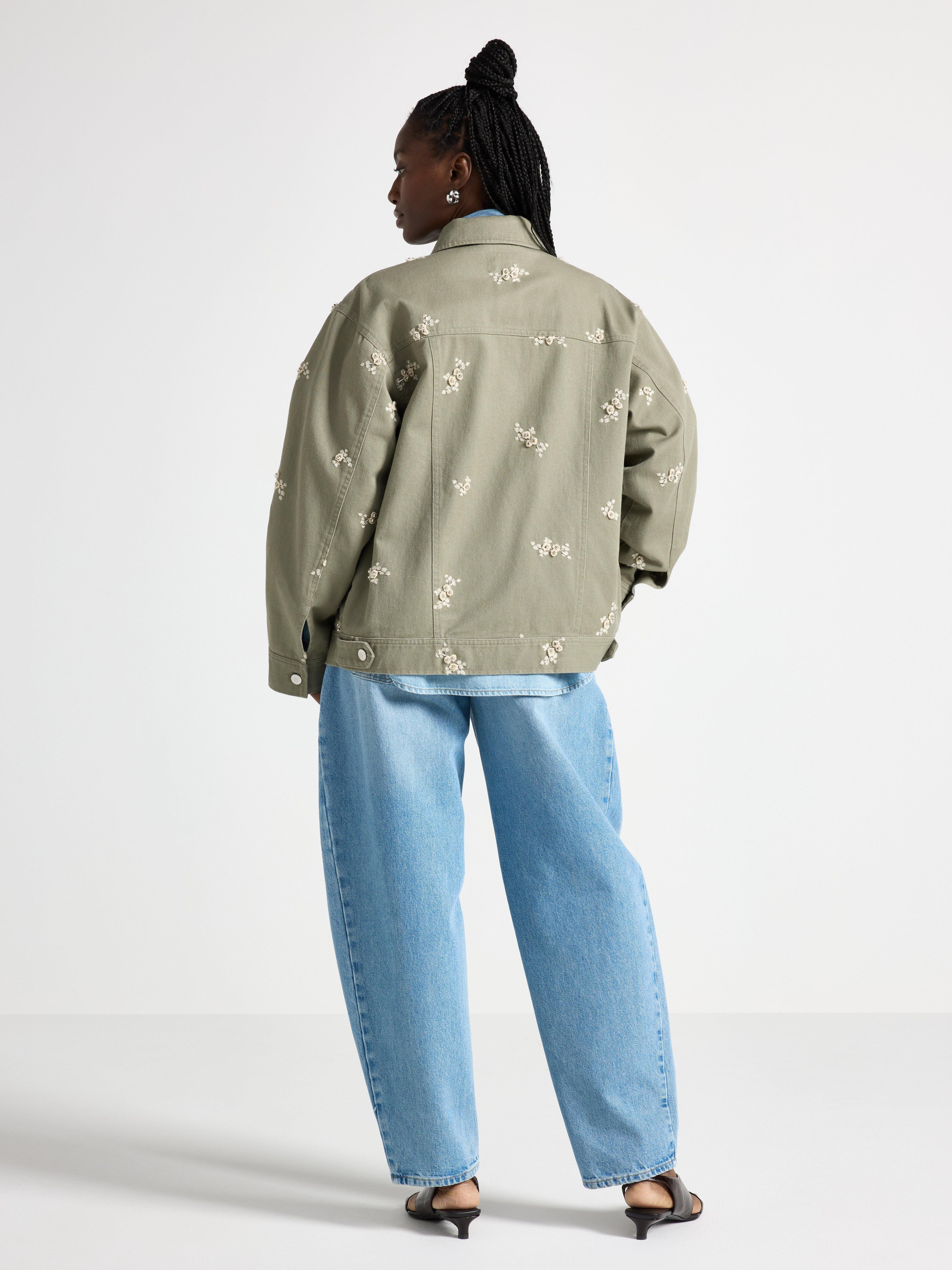 En kvindes ryg i en lys khakifarvet jakke med broderi og et par jeans.