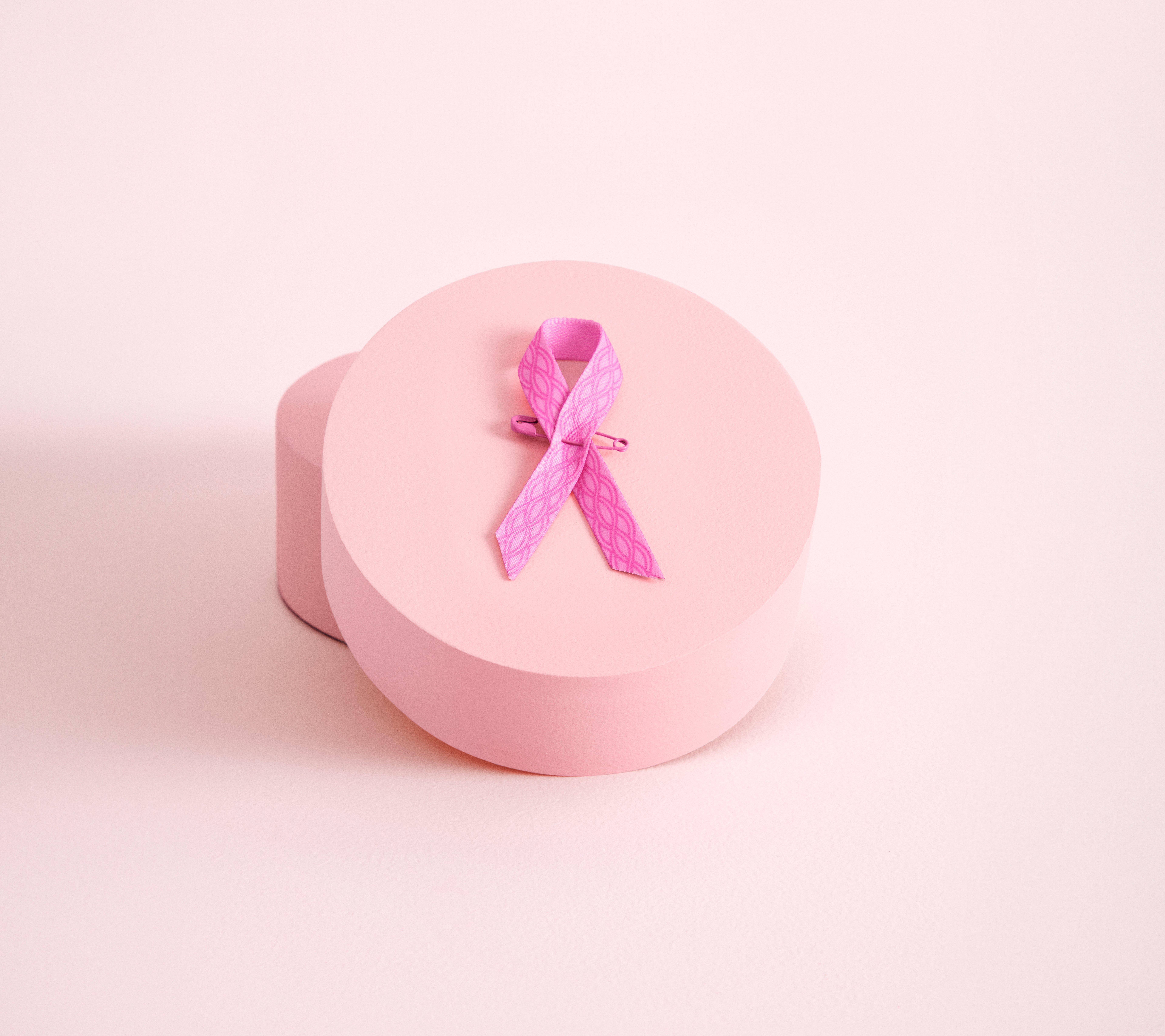 Detailní záběr letošní růžové stužky ležící na kulaté růžové krabičce. Stuha je světle růžová a má tmavší růžový vzor ve tvaru řetězu.