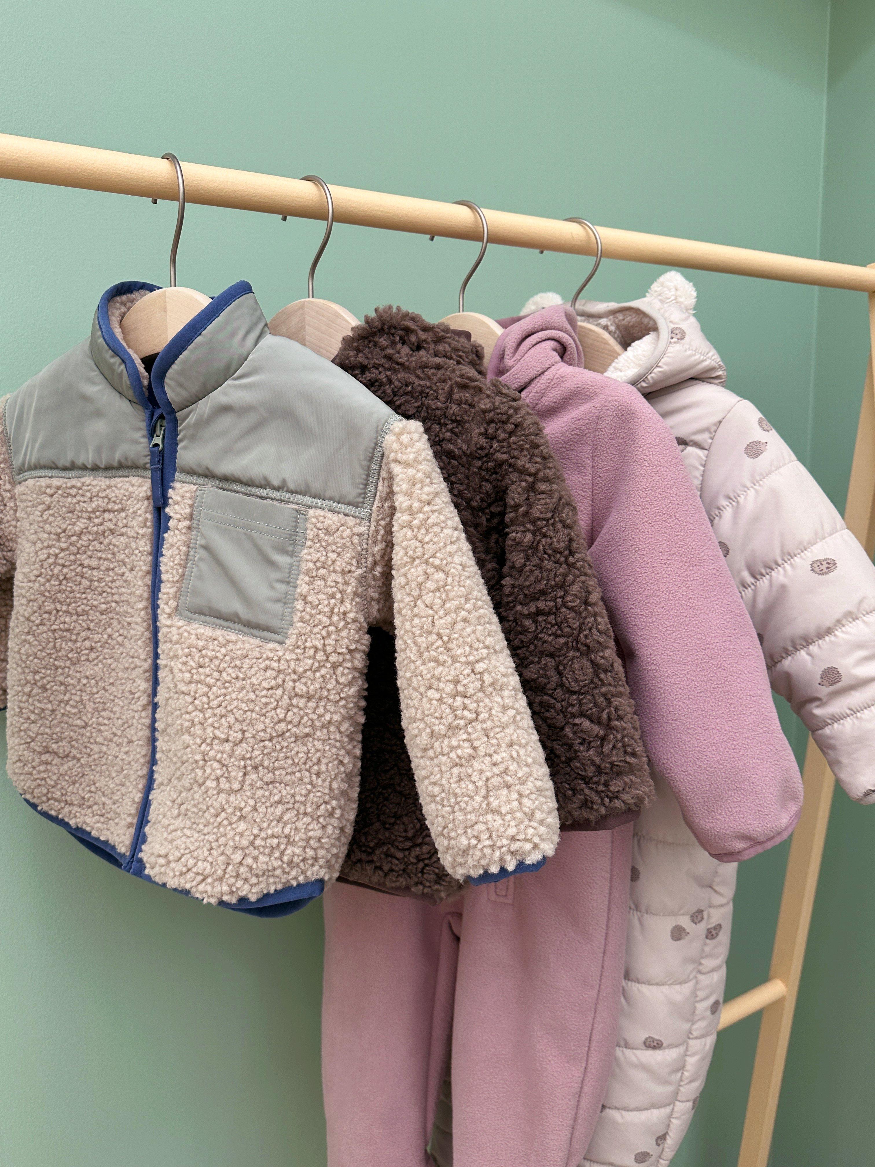 Various outerwear for a baby hangs on a wooden rack.
