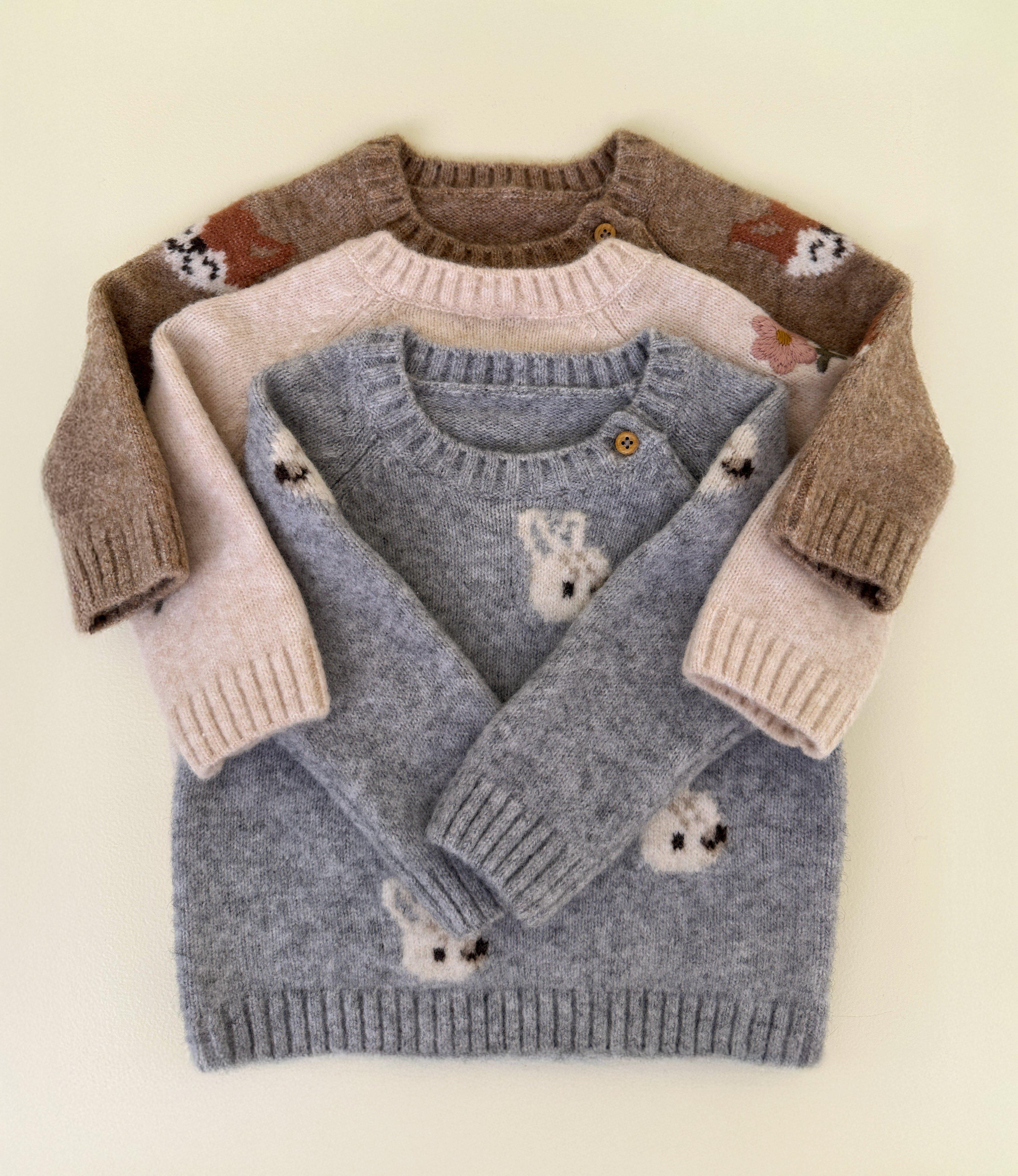 Three knitted baby sweaters. One brown, one pink and one blue-gray, all with animal motifs, lying on top of each other in a pile.