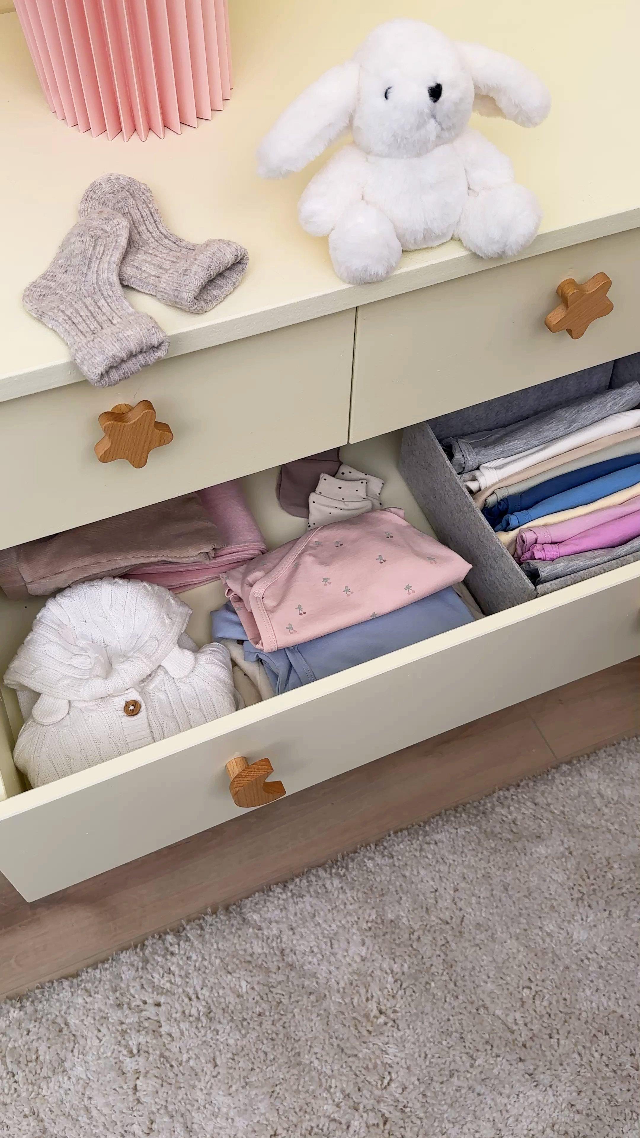  A dresser with an open drawer. Baby clothes are neatly folded in the drawer. On the dresser are a pair of baby socks and a cuddly bunny.