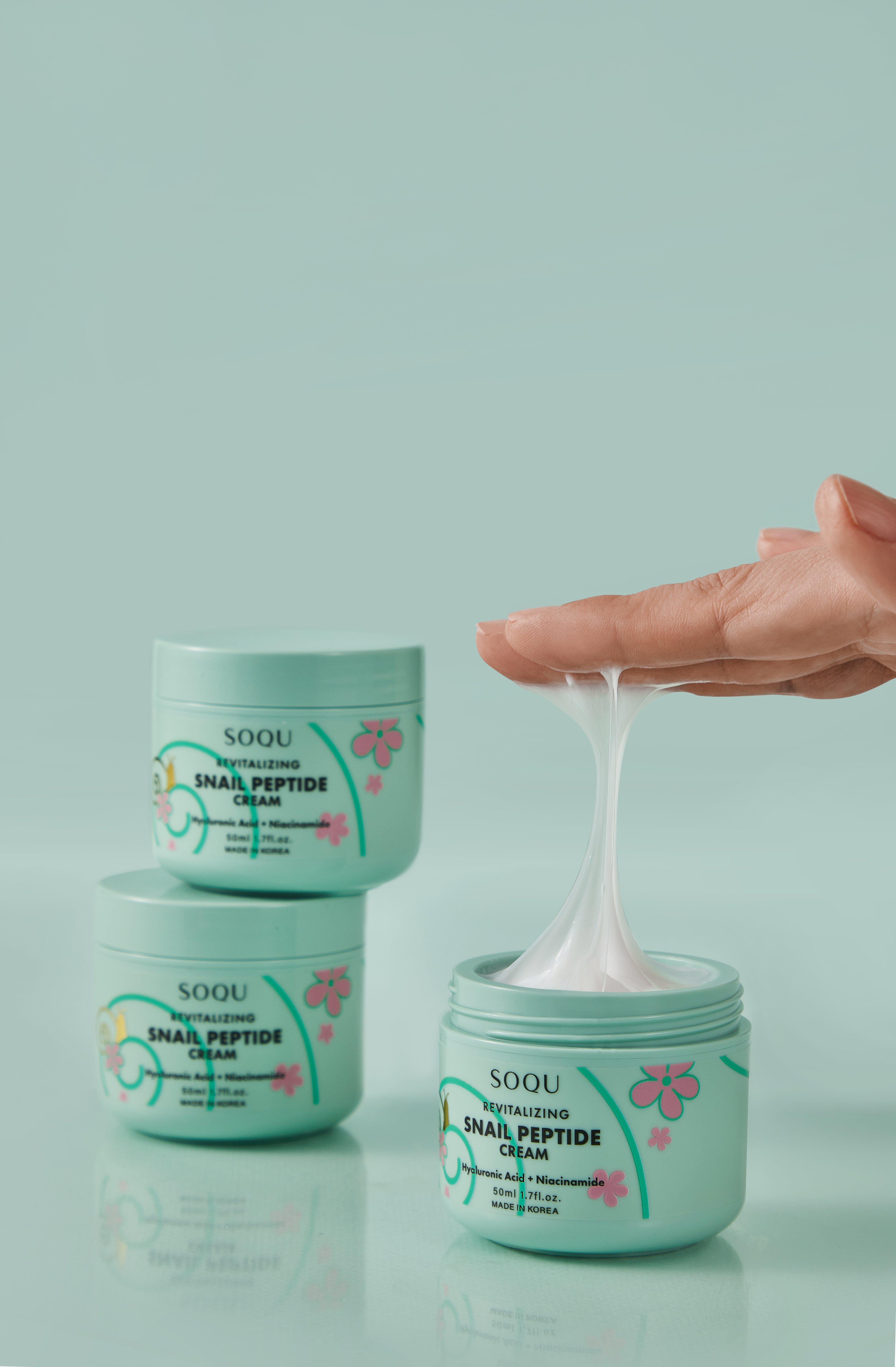 Nærbilde av en hånd som har tatt en dråpe SOQU Snail Peptide ansiktskrem fra en grønn krukke. To bokser til er synlige i bakgrunnen.