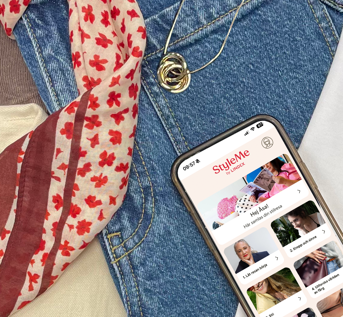 En  iphone som visar startsidan i StyleMe programmet. Mobilen ligger på ett par jeans omgiven av andra klädesplagg.