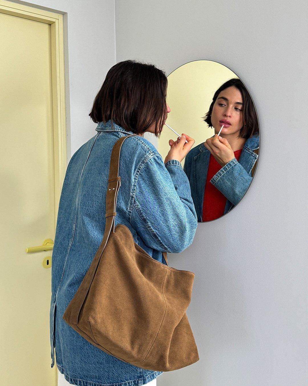 En mörkhårig kvinna klädd i en jeansskjorta tittar sig i en rund spegel och sätter på sig läppstift.