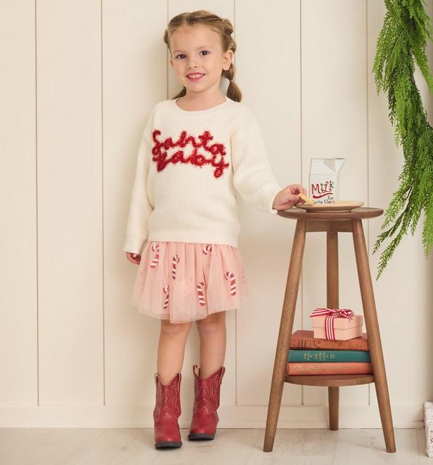 Little girl wearing a candy cane skirt and a white sweater with santa baby saying on it.