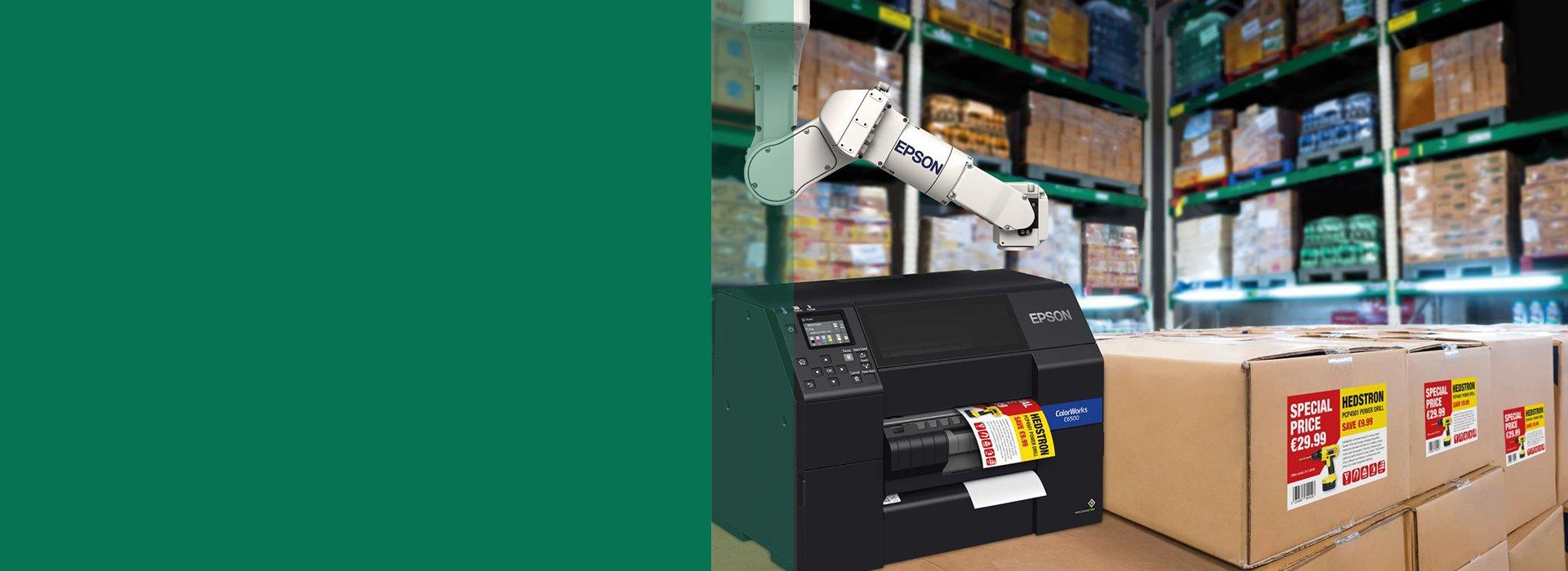 A ColorWorks printer placed next to boxes and a robot arm in the back, all situated in a warehouse