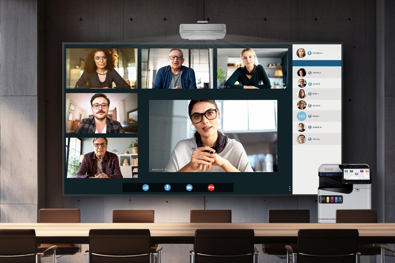 A conference room with a large screen showing a video call. Six participants are visible, each in their own video window.