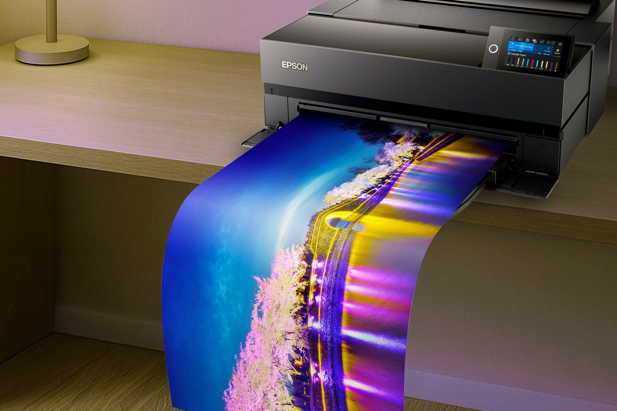 An Epson SureColor printer mid-print. The long, vibrant printout is hanging over the desk.