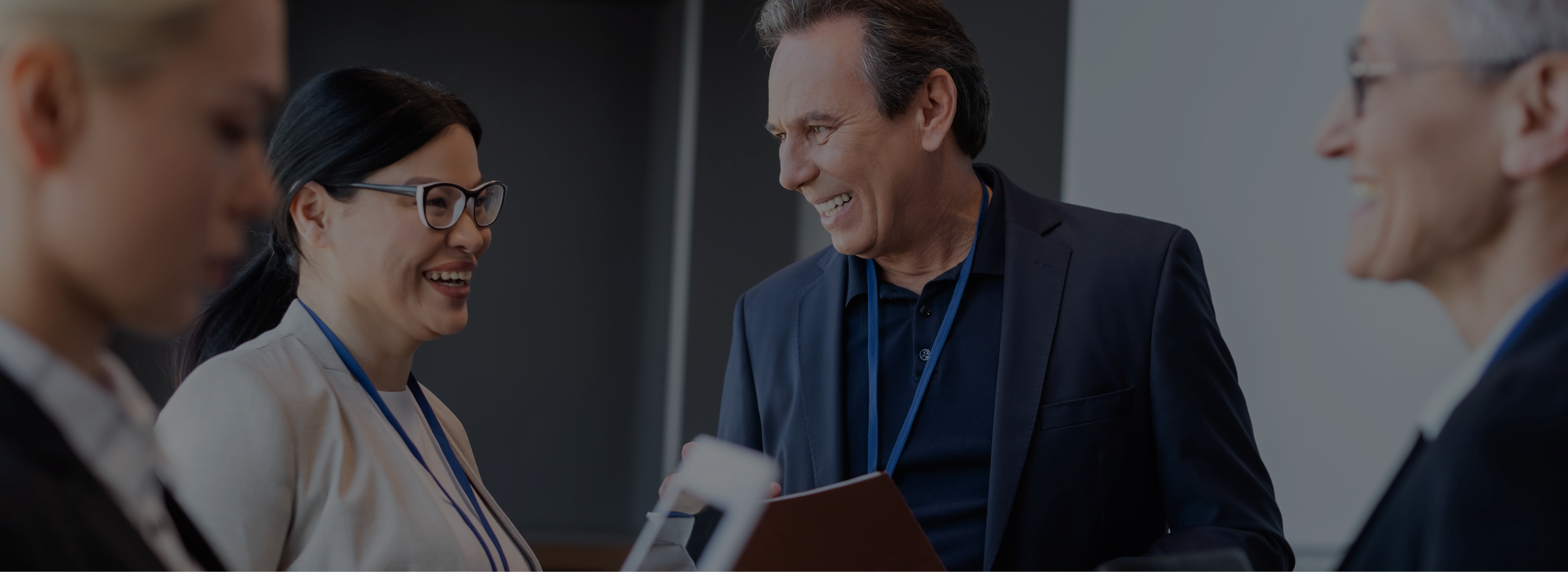 A diverse group of professionals engaged in conversation; a woman smiling, holding documents, and two people with ID badges suggest a positive, collaborative meeting vibe.
