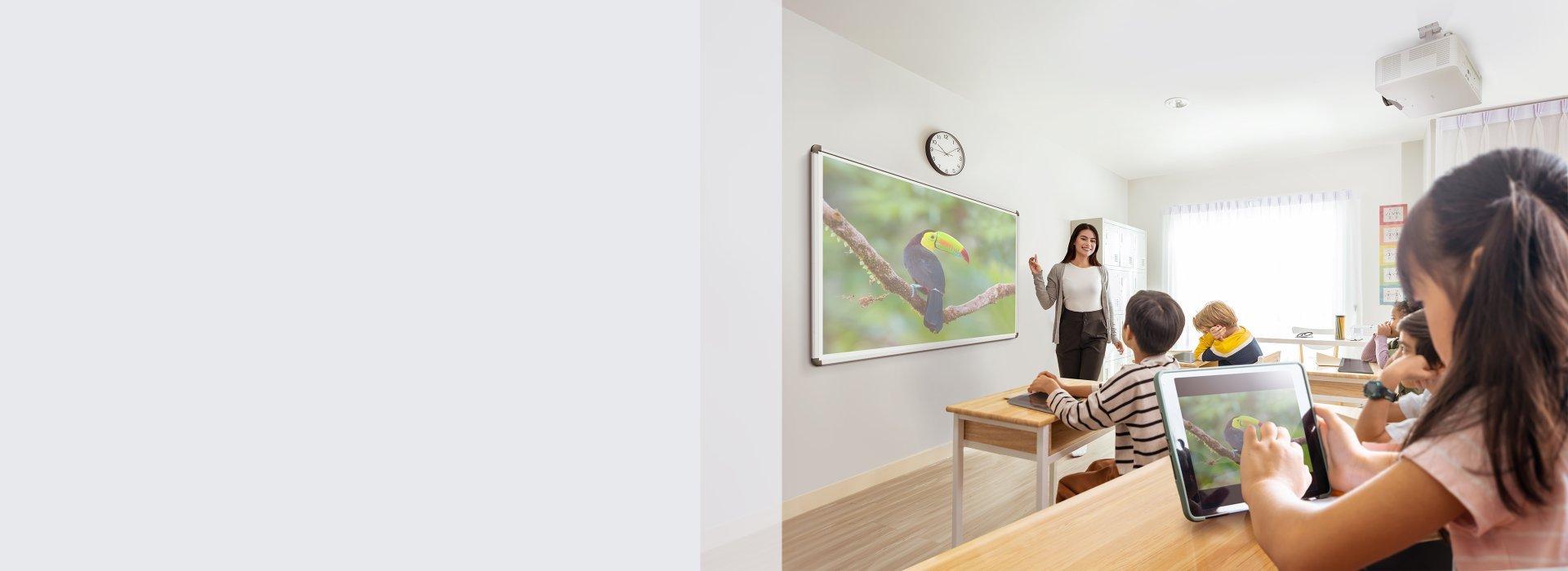 A teacher stands in a bright classroom pointing at a digital board showing a toucan. Students focus on tablets, engaged and attentive.
