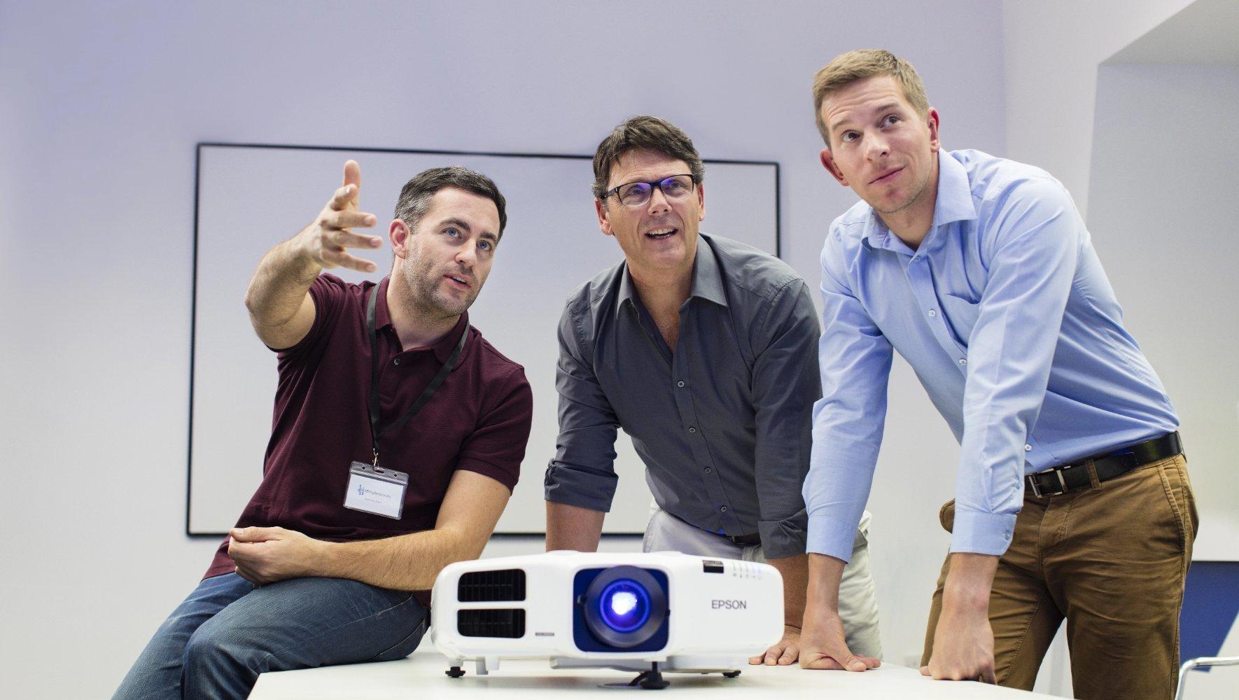 Three men engage in a discussion around a table, looking focused and collaborative. A projector sits on the table, emitting a blue light.