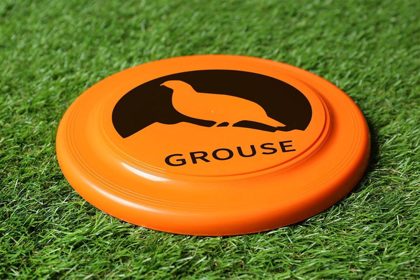 An orange frisbee with a black silhouette of a bird and "GROUSE" text is on green grass.