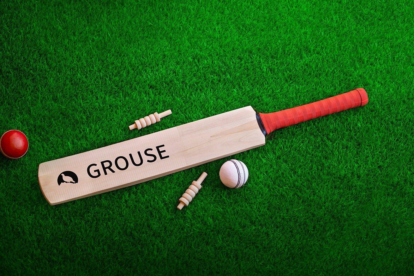 Cricket bat labeled "GROUSE" with red grip lies on green grass.