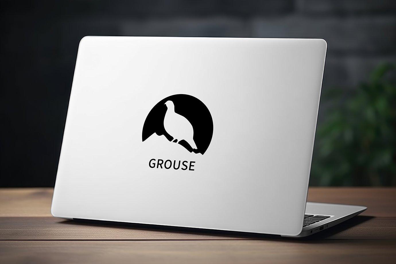 A sleek laptop on a wooden desk displays a black silhouette logo of a grouse within a circle.
