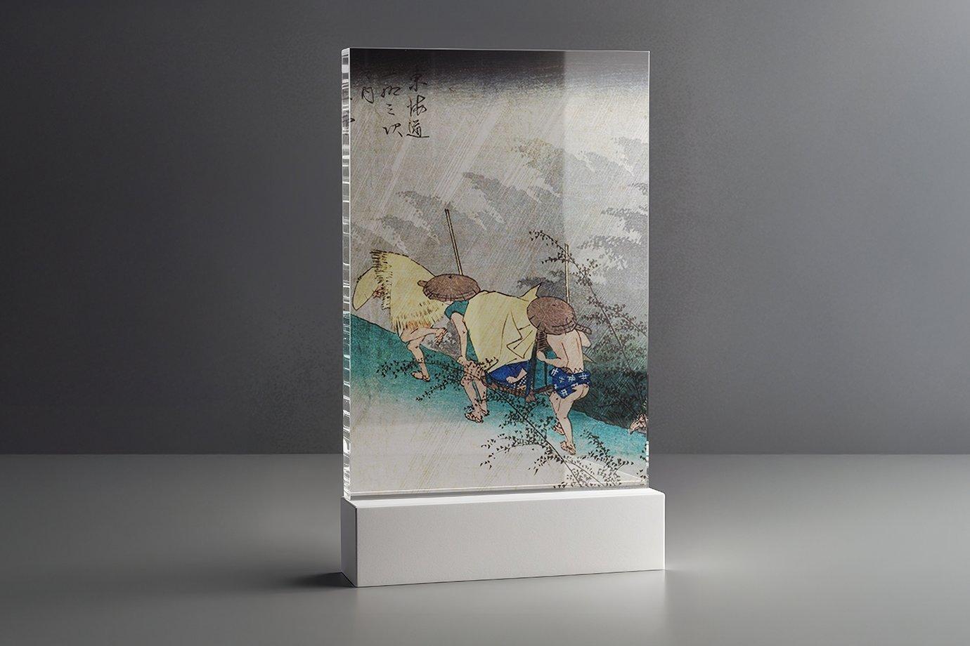 Embossed artwork in an acrylic block depicts two figures in straw hats walking in the rain.