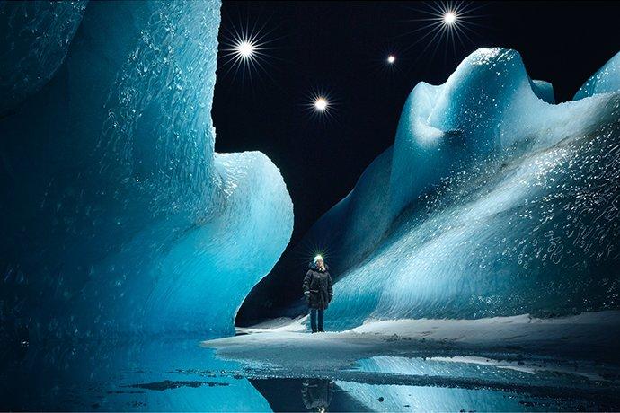 Una persona con cappotto e torcia in testa davanti a ghiacciai illuminati dalla luce delle stelle
