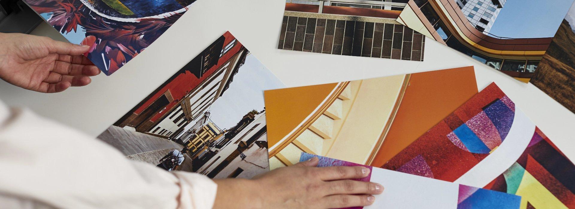 Various vibrant printed photographs are scattered on a table. Someone is picking one of them up for a closer look.