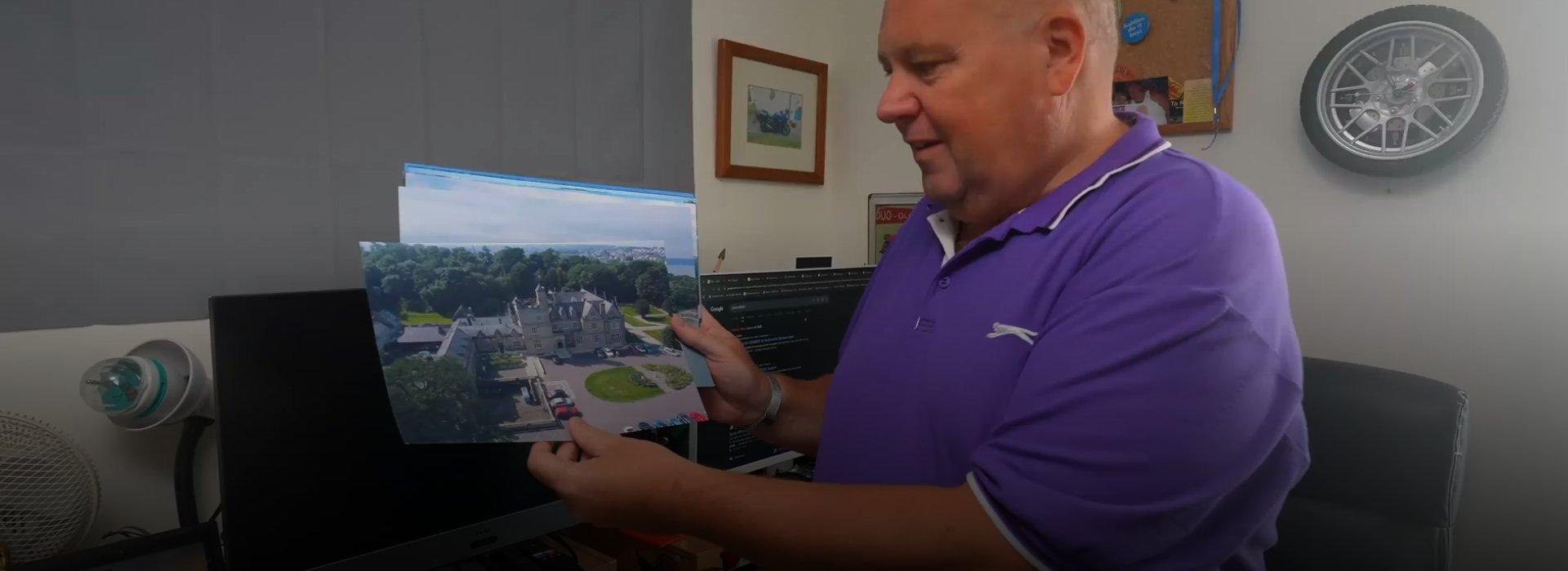 A man in a purple t-shirt holding and showing off various printed aerial photographs.