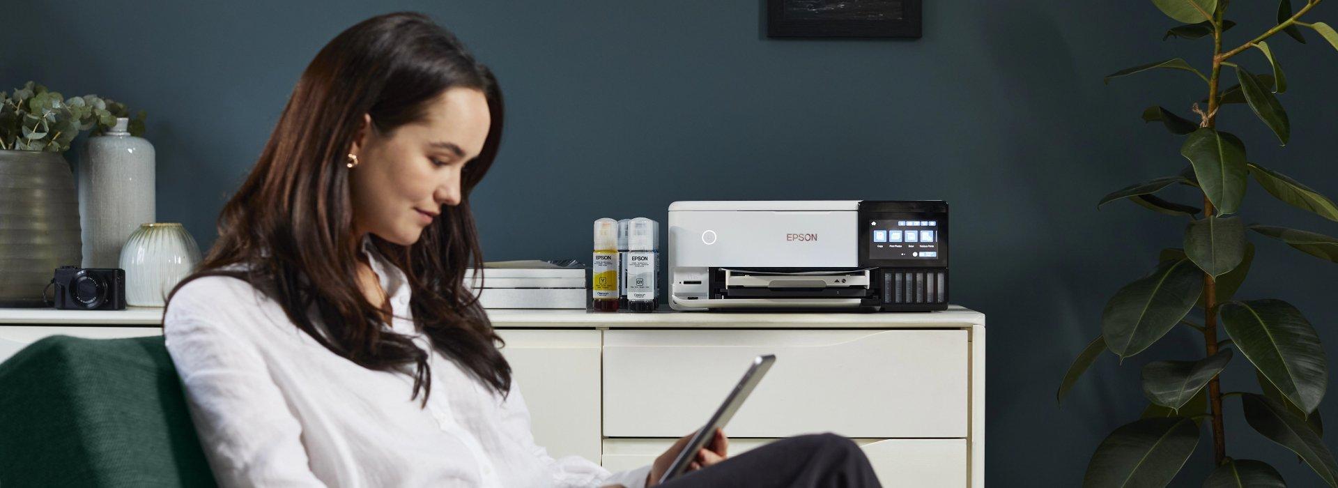 A woman sits on a couch using a tablet in a modern room with a white Epson printer on a dresser with ink bottles beside it. 