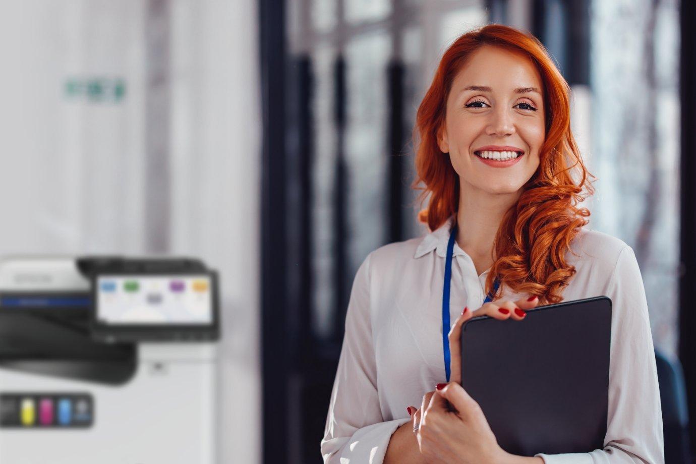 Smiling woman with red hair holding a tablet stands in an office setting. A printer is visible in the background, conveying a professional tone.