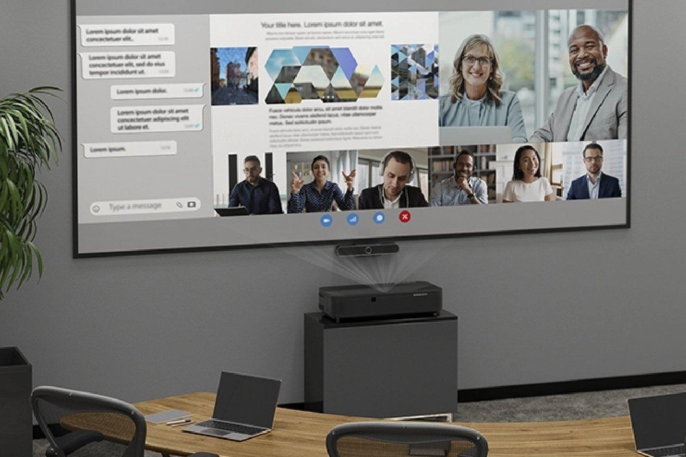Meeting room with a projector displaying a video call on a screen. Six people are visible on the call. Laptops are on a wooden table.