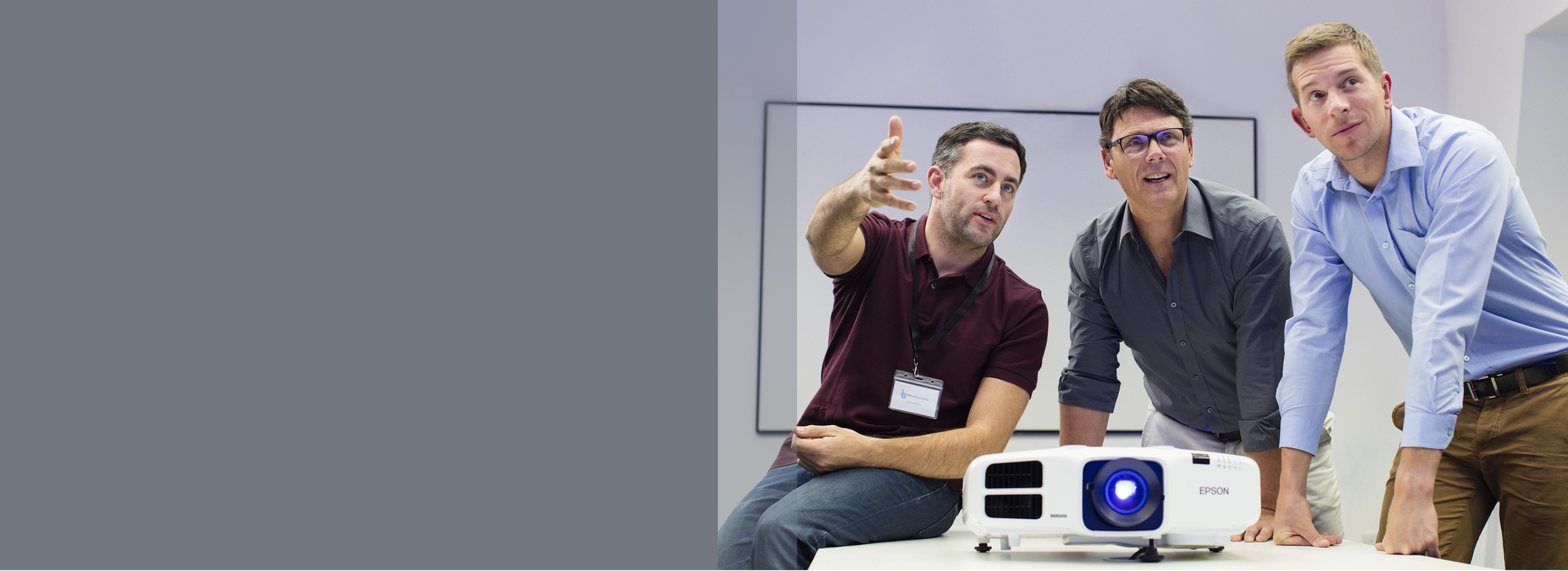 Three men stand around a table with a projector, engaged in discussion. One gestures animatedly. The mood is collaborative and focused.
