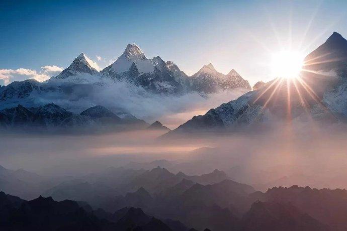 Ein wunderschöner Ausblick auf Berge, die aus den Wolken ragen, einen strahlend blauen Himmel und die Sonne, die über den Bergen hervorkommt.