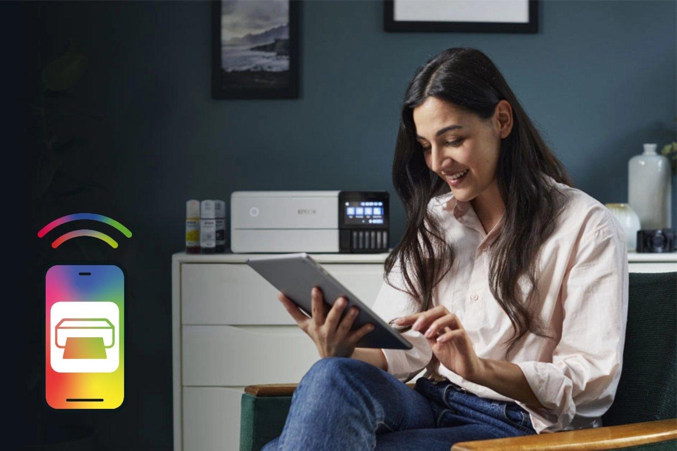 A woman in a cozy room smiles while using a tablet, sitting in front of a printer. A colorful smartphone icon with printing signals is on the left.