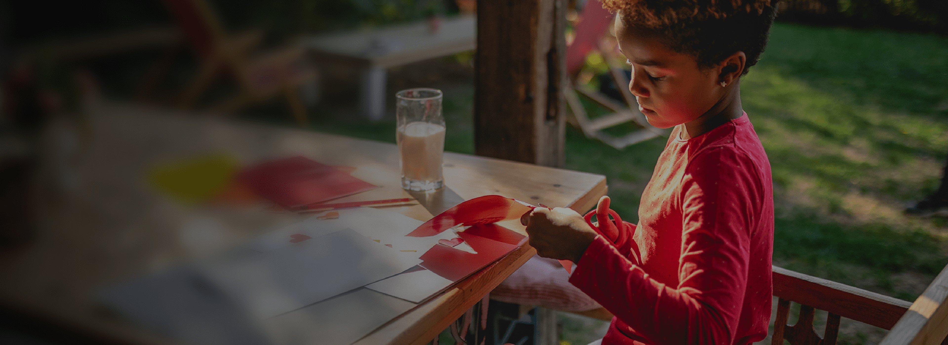 A child in a red shirt sits at an outdoor table, focused on cutting paper for a craft. A glass of milk is nearby. The setting is sunlit and relaxed.