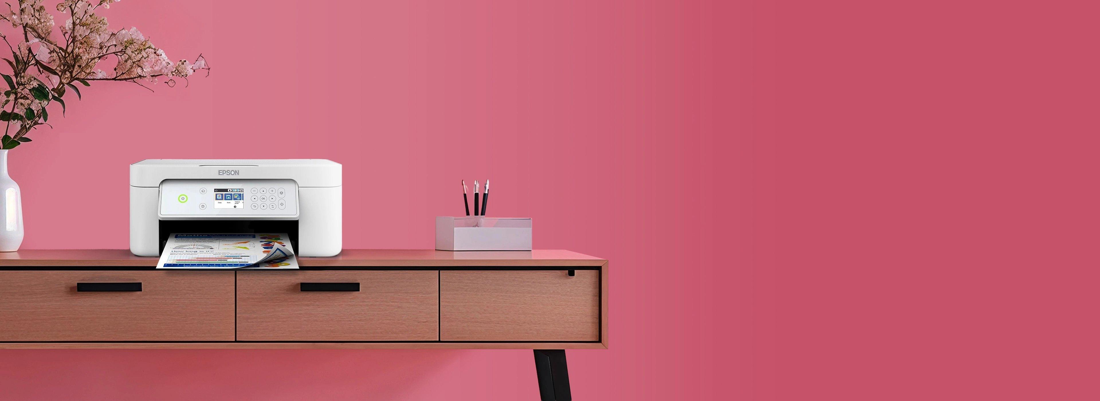 A sleek, white printer on a wooden desk prints colorful paper. A vase with delicate flowers and a box of pens sit nearby, set against a vibrant pink wall.