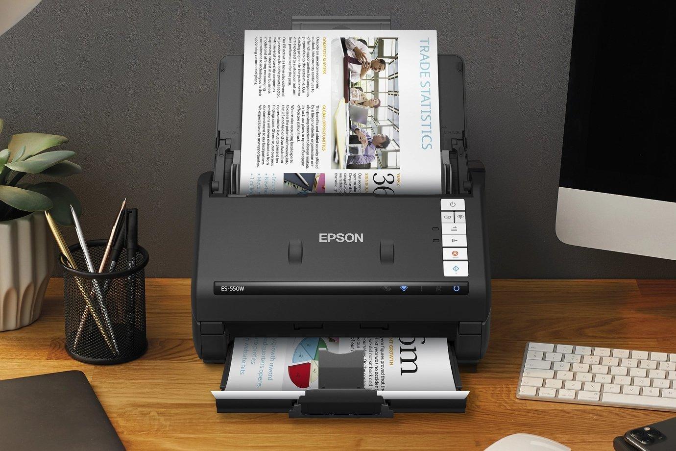 A black Epson scanner on a wooden desk processes a colour document.