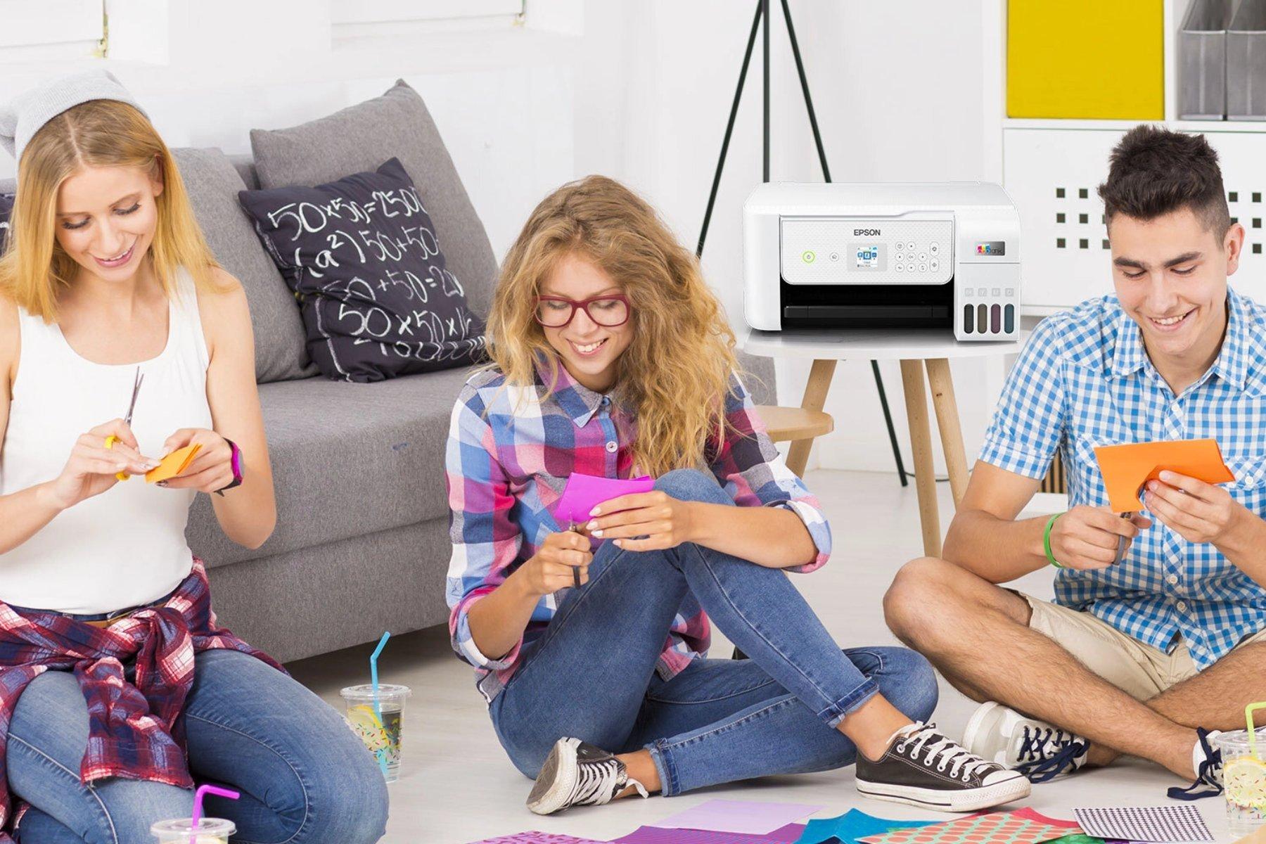 Three people sit on the floor, smiling and crafting colorful paper shapes, with art supplies around. A printer is in the background. The scene is bright and cheerful.
