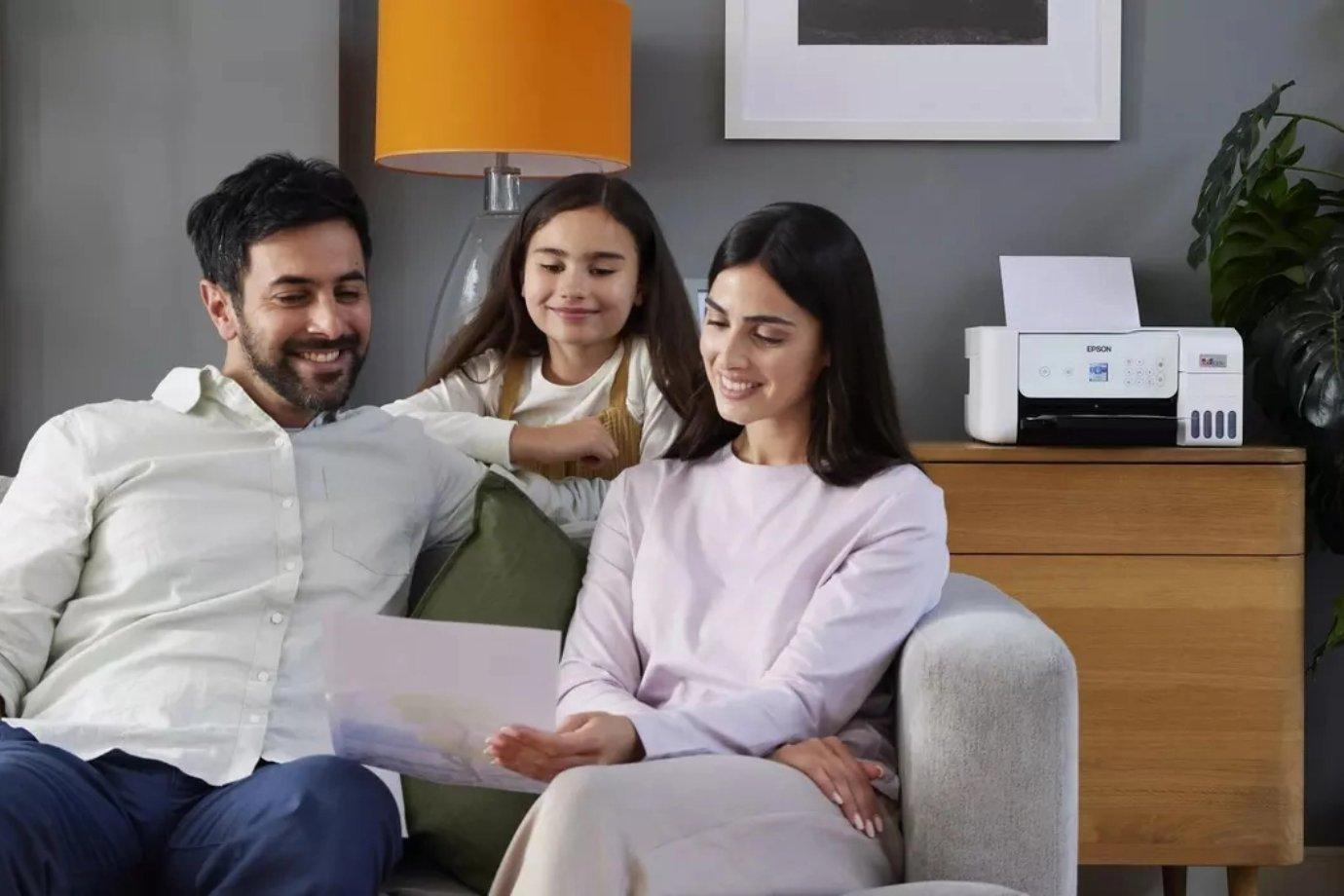 A family of three sits on a sofa, smiling and looking at a printed photo. A printer is on a side table, with a warm lamp and modern decor around them.
