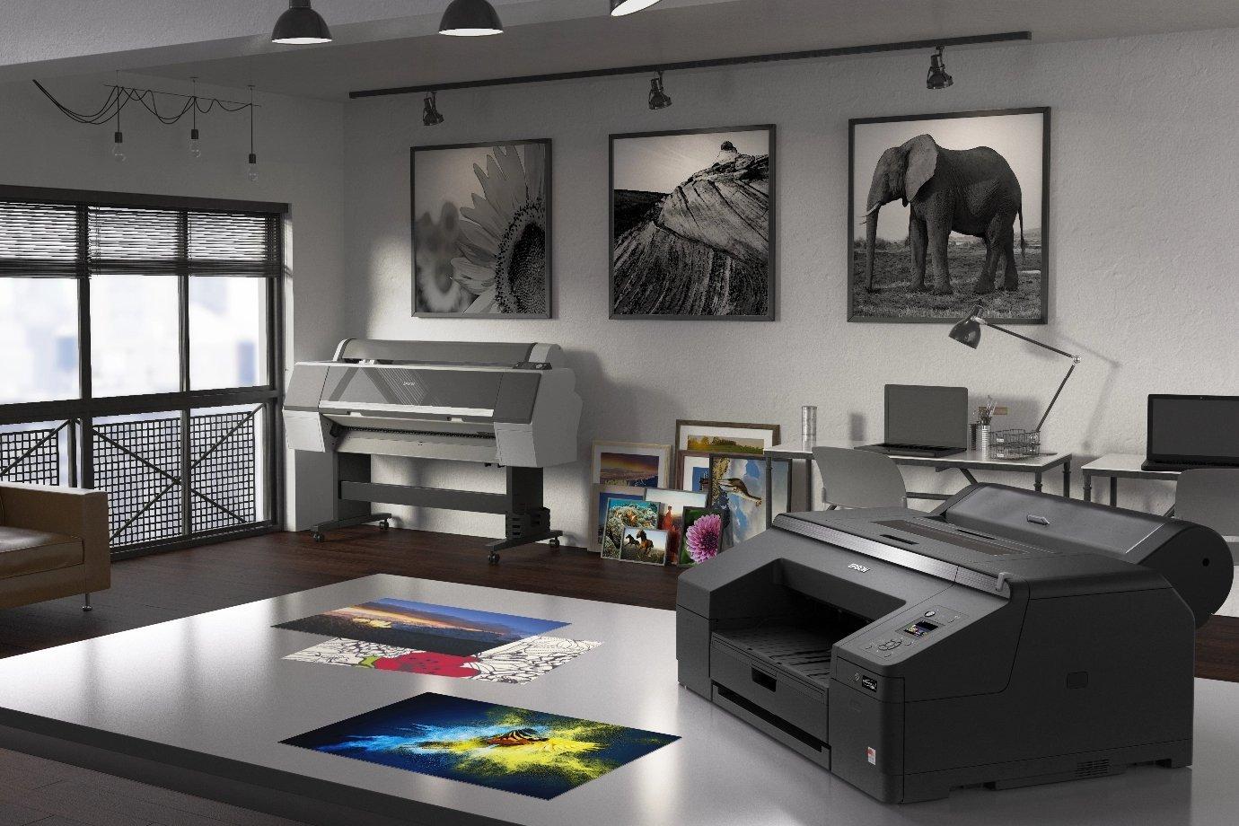 Black Epson printer sat on a desk with printed pictures on a desk, framed images on a wall in the background on a grey wall