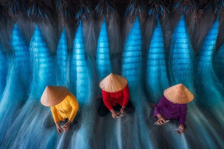 Three people wearing conical hats and colorful clothing sit on blue fishing nets, sorting or weaving, viewed from above, with light streaming through the nets, creating a visually striking pattern.