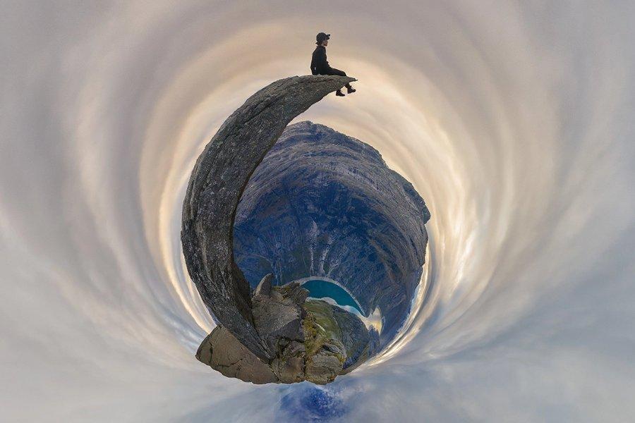 A person sits on the tip of a curved rock ledge high above a mountainous landscape, shown in a surreal tiny-planet panoramic effect that swirls the sky and terrain into a circular vortex.