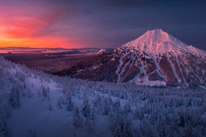 Snow-covered mountain and forest at sunset, with vibrant pink, orange, and purple clouds in the sky and the sun low on the horizon, casting a warm glow over the wintry landscape.