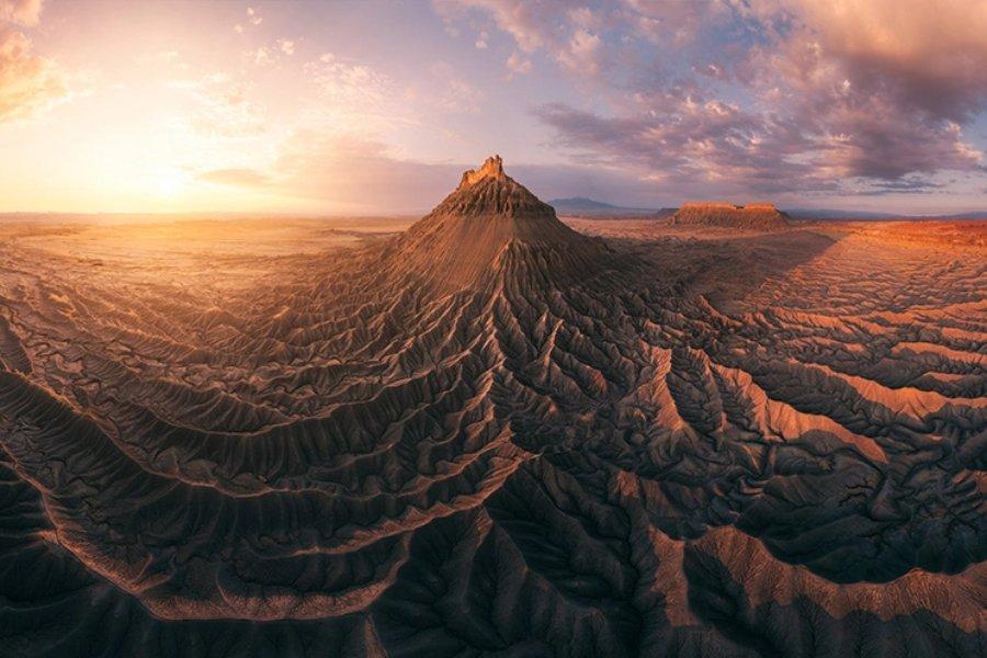 A panoramic view of a rugged desert landscape at sunrise, featuring dramatic ridges, deep shadows, and a central flat-topped mesa under a partly cloudy sky.