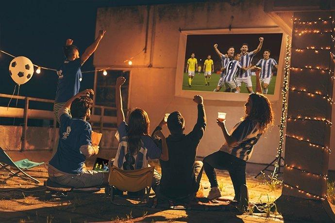 A group of people cheering watching a football game on a projection screen at night outside, seemingly on a rooftop seating area.