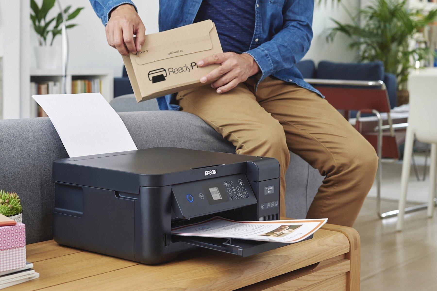 A person inserts a paper into a black Epson printer on a wooden table. The setting is a home office with bookshelves. The mood is efficient and organized.