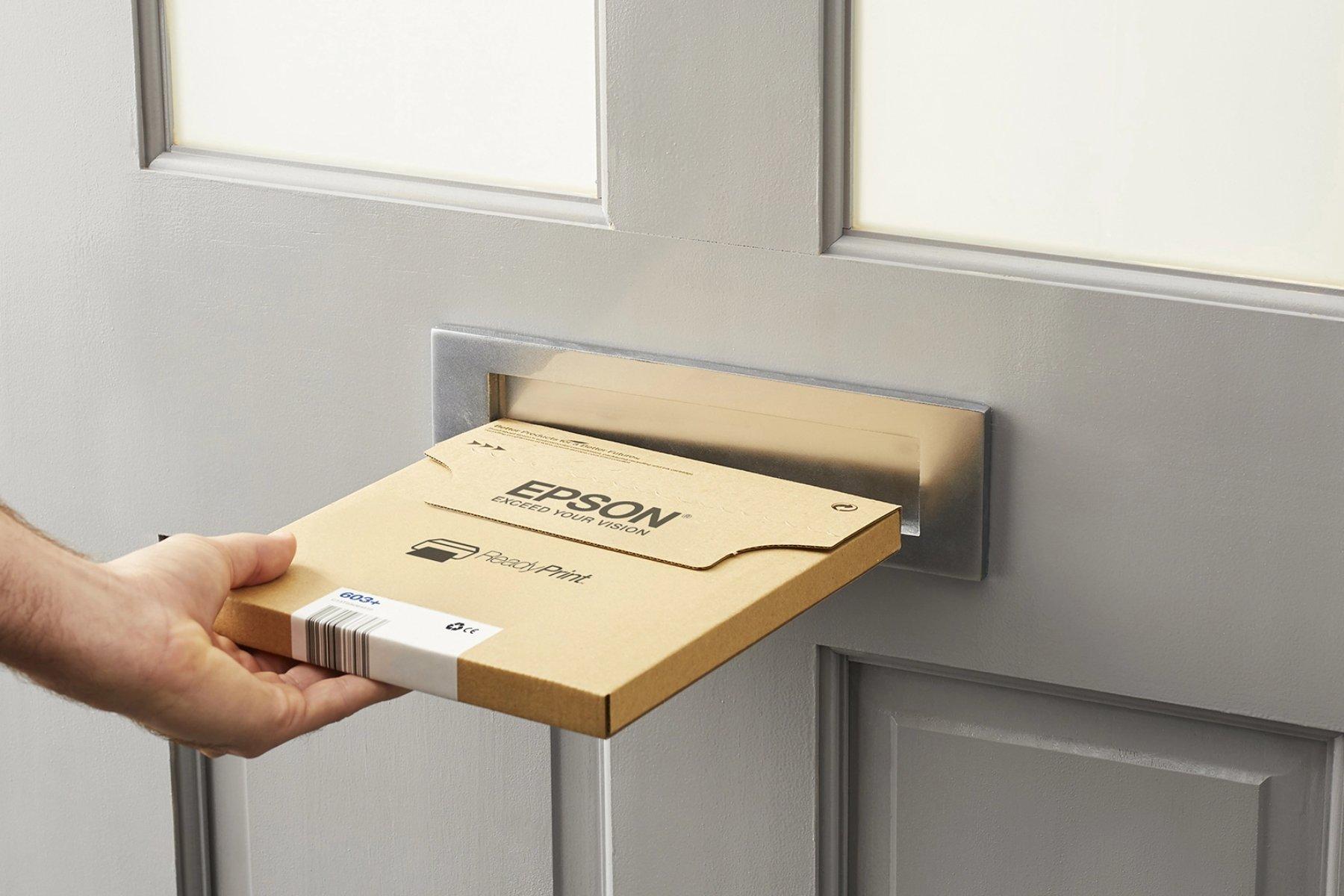 A hand inserts a flat, rectangular Epson cardboard package into a silver mail slot in a light grey door, conveying convenience and efficiency.