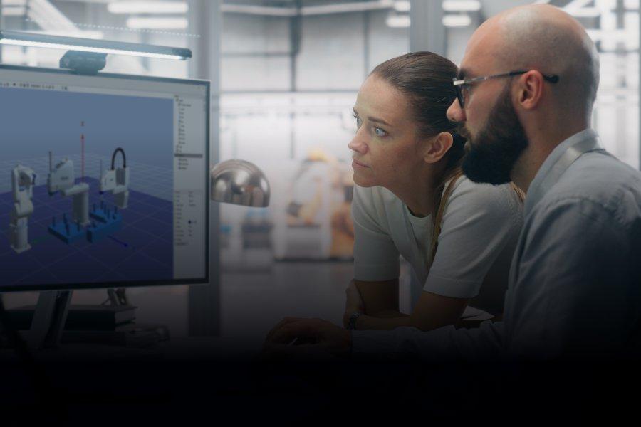 A man and a woman in a factory setting, looking at a screen with Robot Software open - displaying 3D models of the robots.