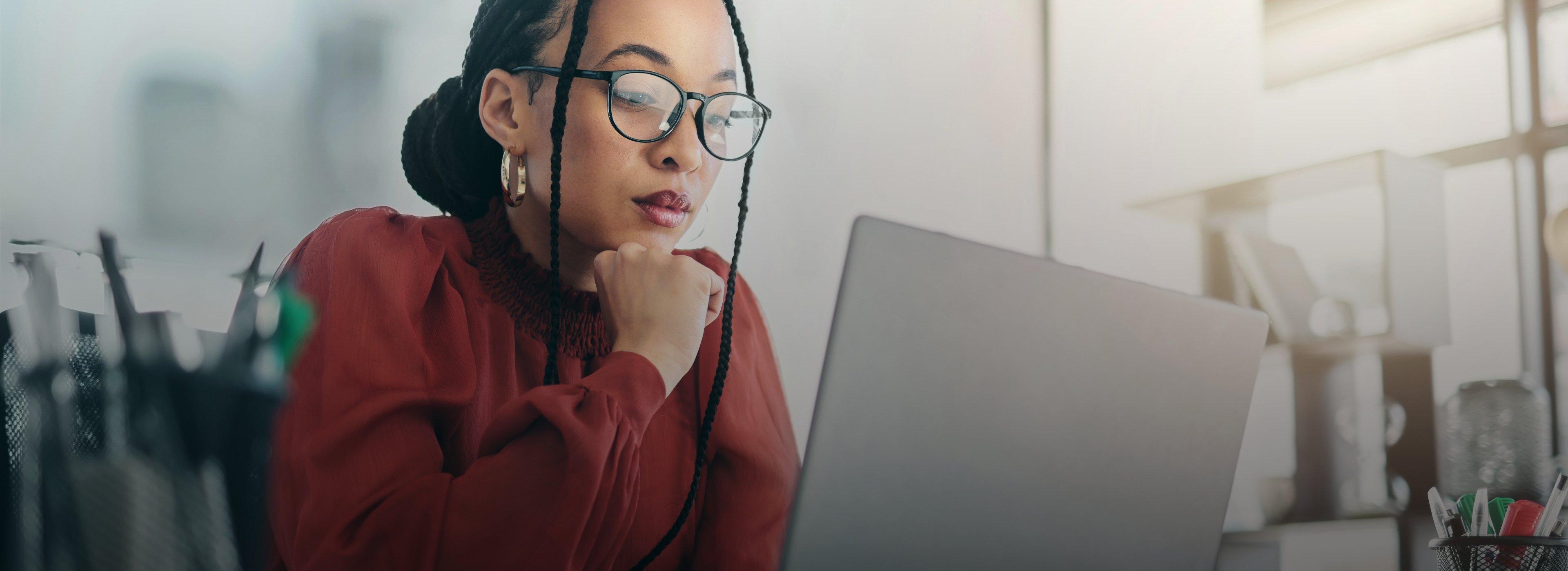 A woman in glasses and a red blouse works intently on a laptop in a bright office.