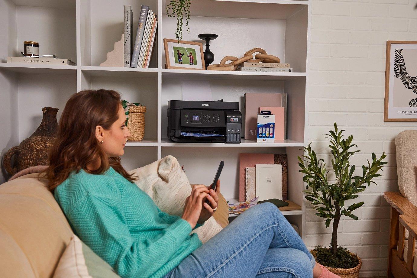 A woman in a teal sweater uses a smartphone on a beige couch. Behind her is a white shelf with books, decor, and a printer.