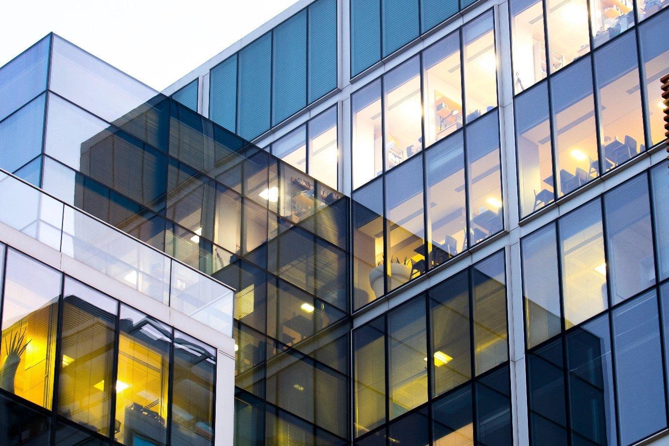 Modern glass office building at dusk, with warm interior lights illuminating desks and chairs.