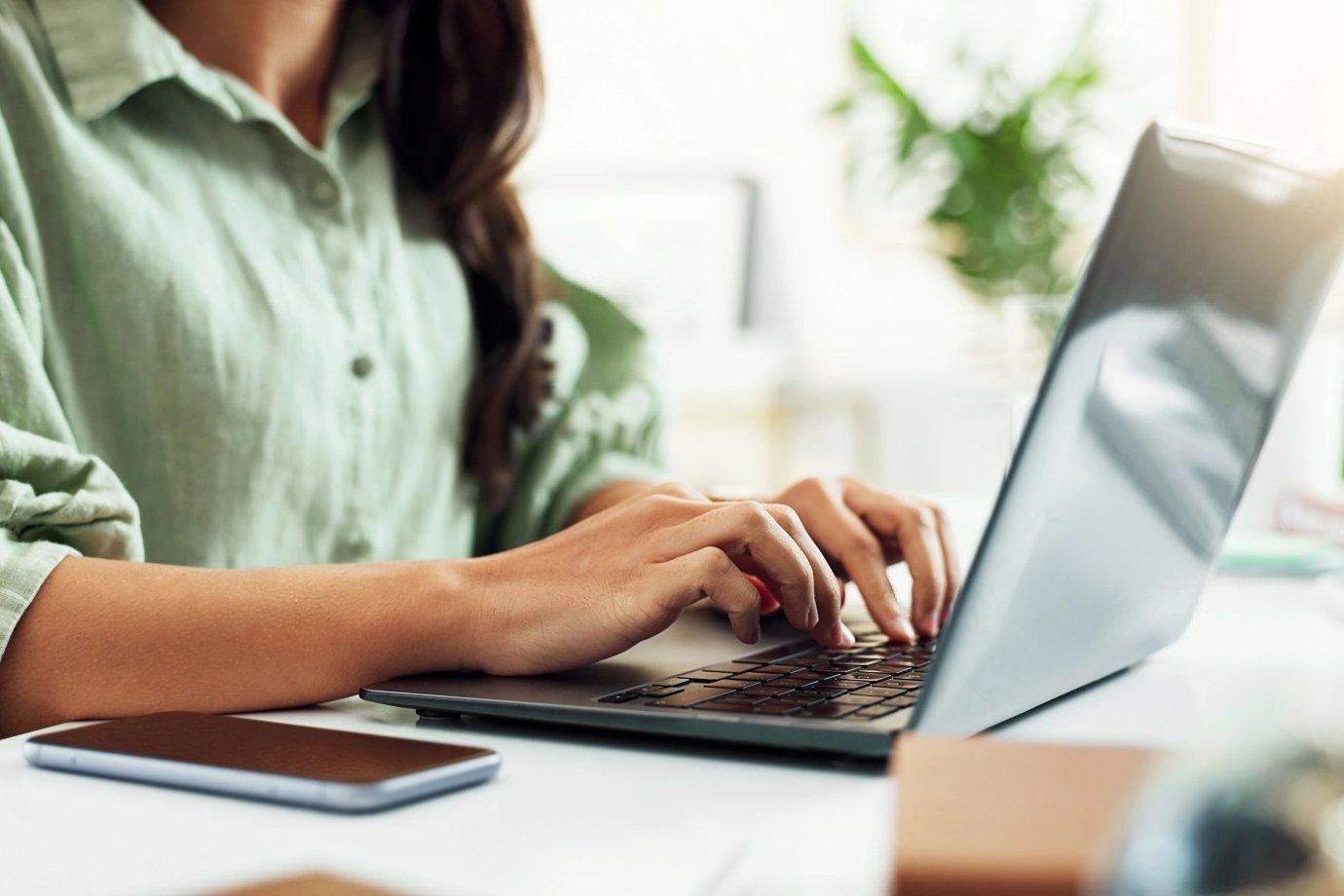 A person in a light green shirt types on a laptop at a bright, sunlit desk.