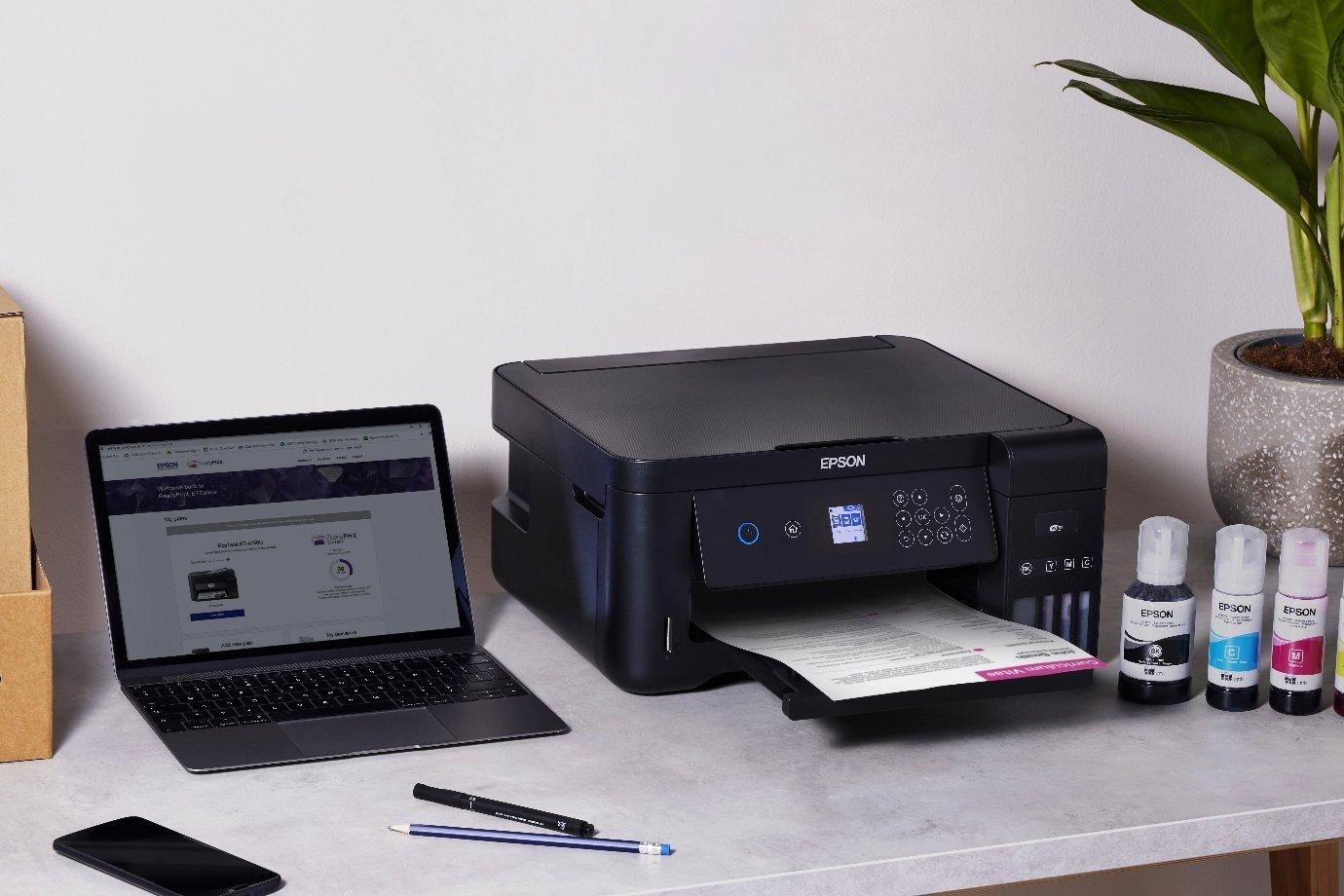 A home office setup featuring a black Epson printer with printed paper, ink bottles & a laptop displaying the Epson website.