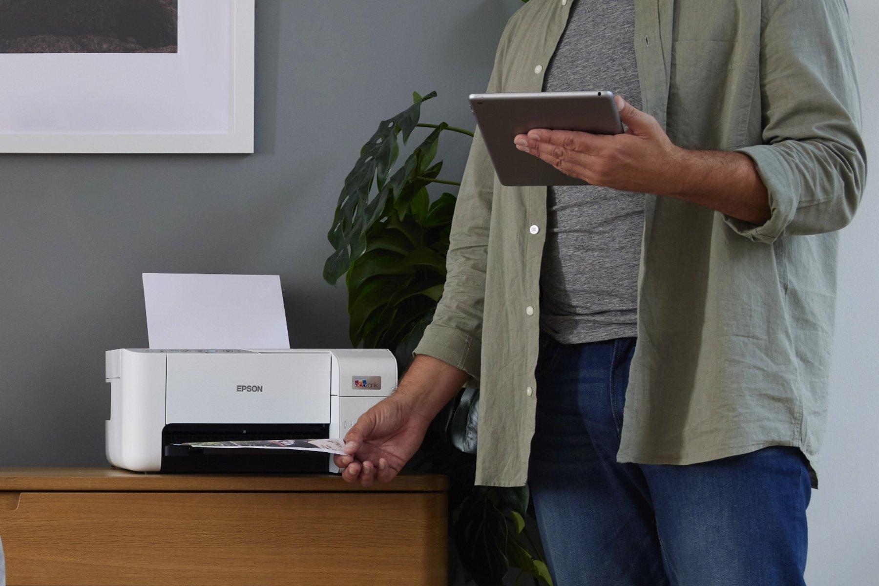 A person, wearing a gray shirt and olive jacket, uses a tablet while standing beside an Epson printer.